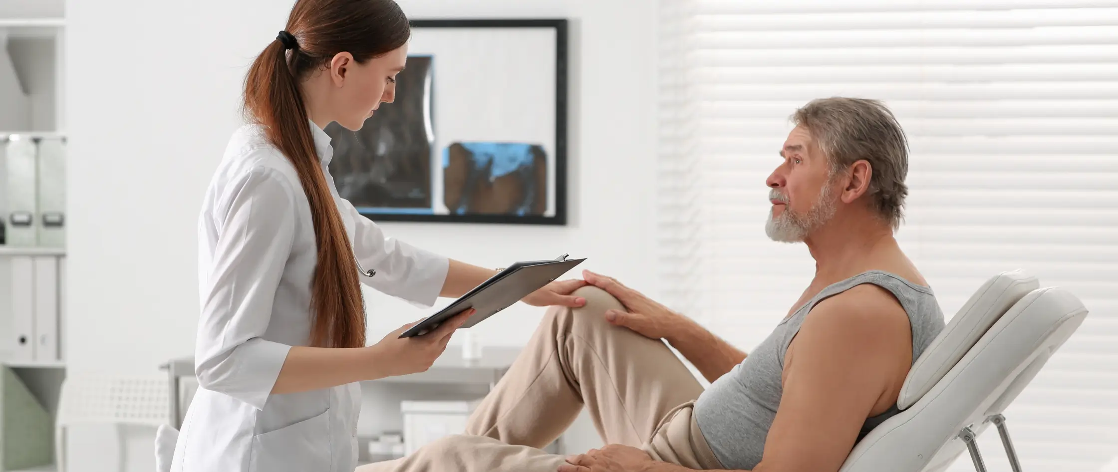 A doctor examines the knee of an elderly man during a medical consultation in a clinical setting.