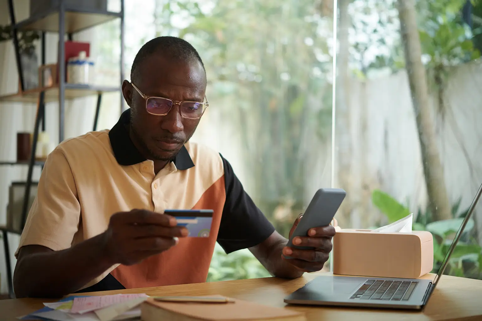 A man with glasses is engaged with his laptop, holding a credit card for online transactions.
